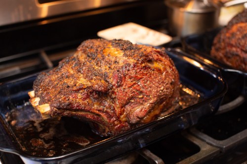 A large, roasted beef joint with a crispy, seasoned crust resting in a dark roasting pan on a stovetop.