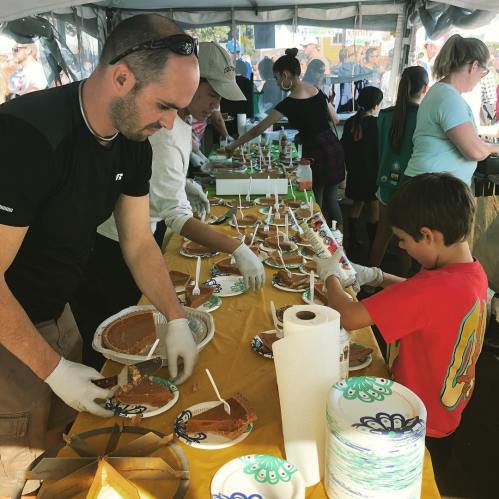 A group of people serving food at a festival, with adults and children preparing plates under a tent.