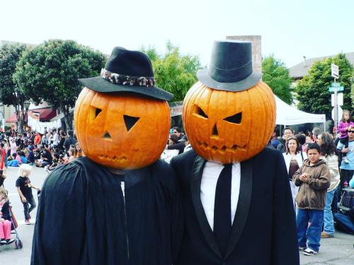 Two people wearing carved pumpkin heads, one in a black cape and the other in a suit, at a festive outdoor event.