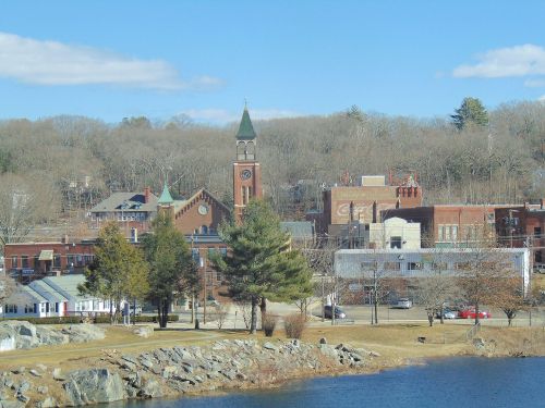 A scenic view of a small town with a clock tower, trees, and a lake under a clear blue sky.
