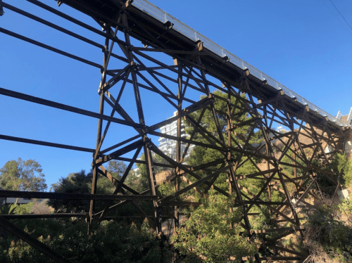 A large, rusted metal bridge structure with a clear blue sky in the background and greenery below.