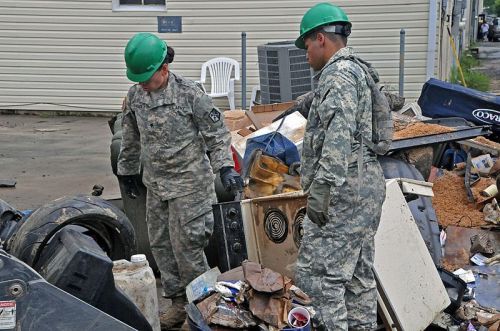 Two soldiers in green helmets inspect a pile of debris and trash in a military uniform, surrounded by scattered items.