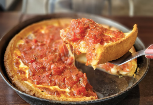 A slice of deep-dish pizza being lifted from a pan, topped with tomato sauce and herbs.