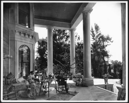 A vintage black-and-white photo of a porch with people sitting, a dog, and lush greenery in the background.