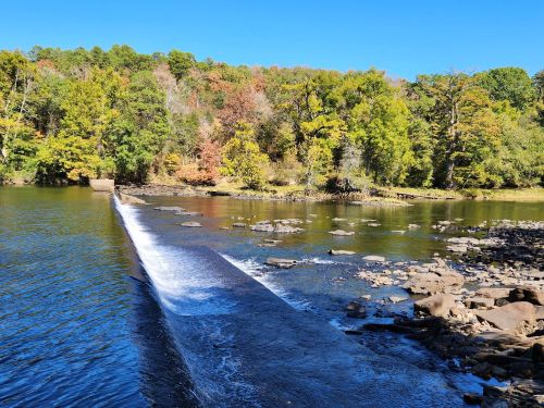 A serene river scene with a small waterfall, surrounded by colorful autumn trees and clear blue skies.