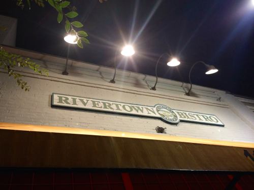 Sign for Rivertown Bistro illuminated at night, mounted on a brick wall with overhead lights.