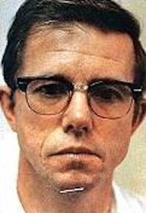 A close-up portrait of a man with glasses, short dark hair, and a serious expression.