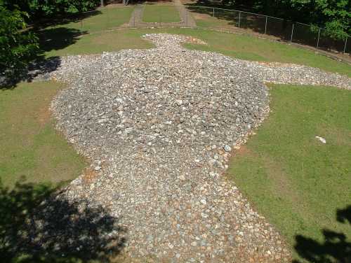 A large stone sculpture resembling a figure, set in a grassy area with trees and a pathway in the background.