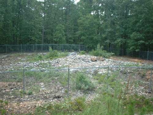 A fenced area with a pile of rubble and sparse vegetation, surrounded by trees in a wooded setting.