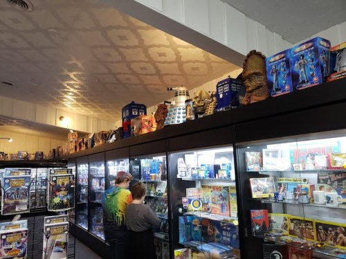 Two people browse collectibles in a store, with shelves displaying various toys and figures, including a Dalek and TARDIS.