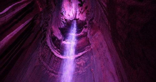 A vibrant purple-lit cave with a waterfall cascading down from the ceiling, creating a mystical atmosphere.