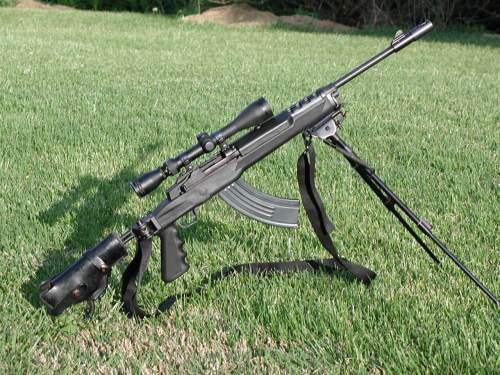 A black rifle with a scope, bipod, and magazine, resting on green grass.