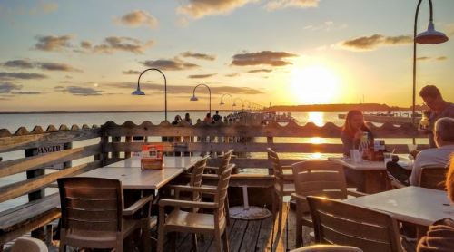 Sunset over a pier with tables and chairs, people enjoying the view, and soft clouds in the sky.