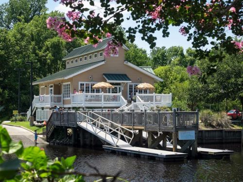 A charming riverside building with a deck, surrounded by greenery and flowers, featuring outdoor seating under umbrellas.