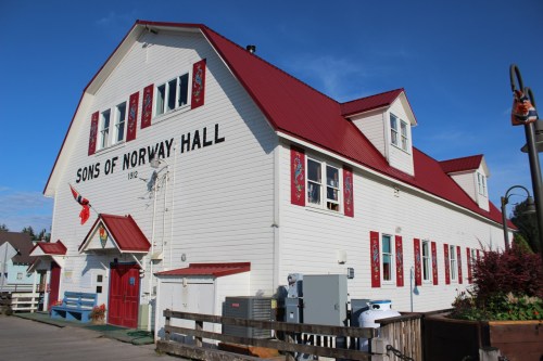 A large white building with a red roof, labeled "Sons of Norway Hall," featuring decorative windows and a clear blue sky.