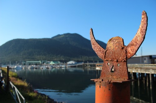 A rusty post with horn-like features stands by a calm harbor, with mountains in the background under a clear blue sky.
