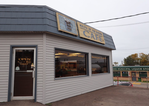 Exterior of "South of the Border Cafe," featuring a sign and open door, with a cloudy sky in the background.