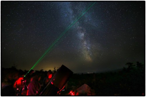 A group of stargazers uses a green laser pointer to highlight the Milky Way in a dark night sky.