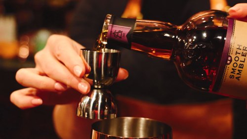 A hand pours liquor from a bottle into a jigger for cocktail preparation.