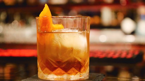 A glass of cocktail with a large ice cube and an orange twist garnish, set on a bar counter.