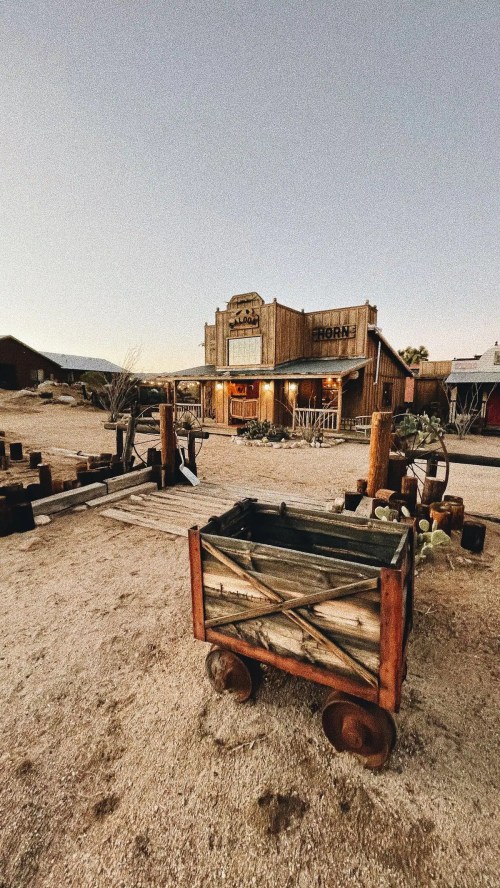 A rustic wooden building with a sign, surrounded by a dusty landscape and an old cart in the foreground.