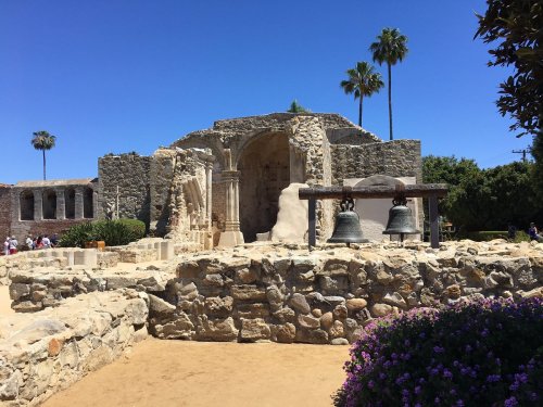 Historic stone ruins with two bells, surrounded by palm trees and vibrant flowers under a clear blue sky.