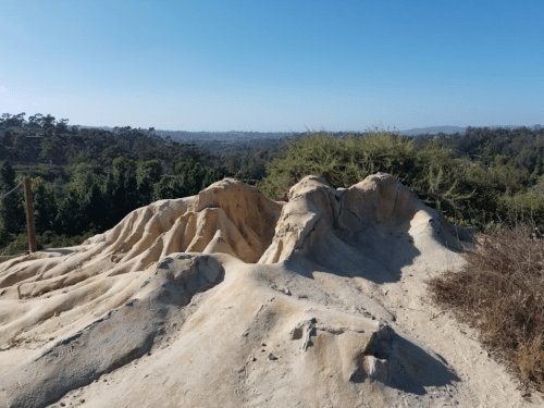 A sandy, textured hill with gentle slopes, surrounded by greenery and a clear blue sky in the background.