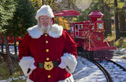 A smiling Santa Claus in a red suit stands in front of a vintage red train surrounded by trees and snow.
