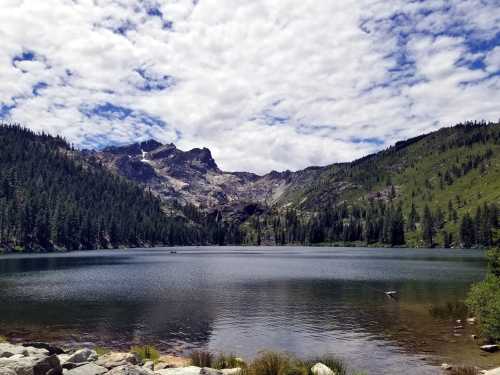 A serene lake surrounded by lush green mountains and a cloudy blue sky, reflecting the landscape in the water.