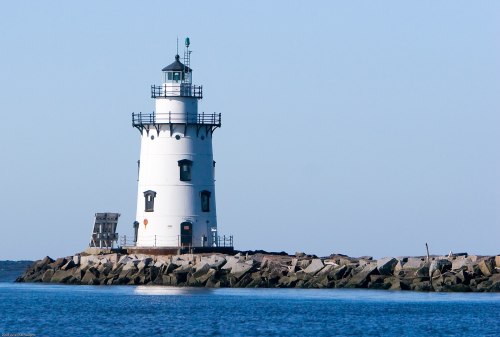 A white lighthouse stands on a rocky jetty, surrounded by calm blue waters under a clear sky.