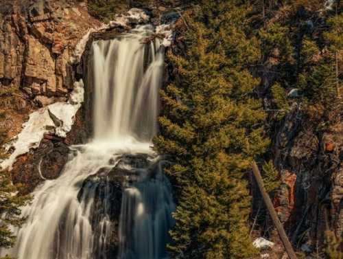 A cascading waterfall flows down rocky cliffs, surrounded by evergreen trees and patches of snow.