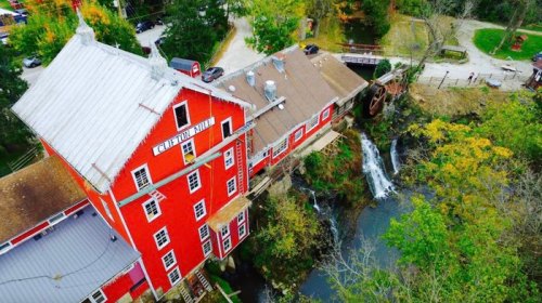 Aerial view of a red barn-like building near a waterfall and lush greenery, with a winding path and vehicles nearby.
