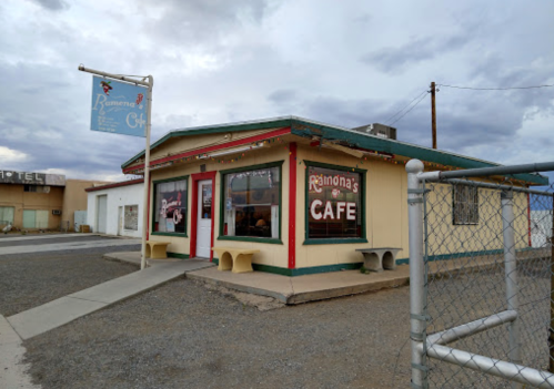 A small, colorful café with a sign reading "Ramona's Café," set against a cloudy sky and gravel lot.