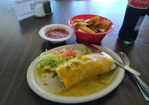 A plate with a cheese-covered burrito, lettuce, and tomato, alongside a bowl of tortilla chips and salsa, with a soda.
