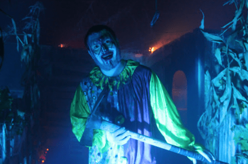 A creepy clown in a colorful costume stands in a dimly lit, foggy setting, holding a weapon and grinning menacingly.