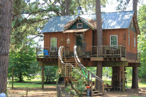 A whimsical treehouse with a wooden exterior, metal roof, and a spiral staircase, surrounded by trees and greenery.
