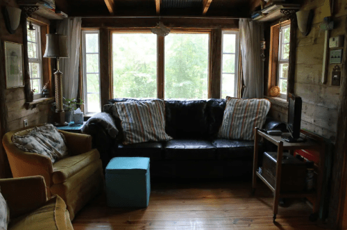 Cozy living room with a black couch, yellow armchair, blue ottoman, and large windows overlooking greenery.