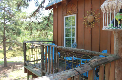 A wooden cabin with a sun decoration, featuring a balcony with blue chairs and surrounded by trees.