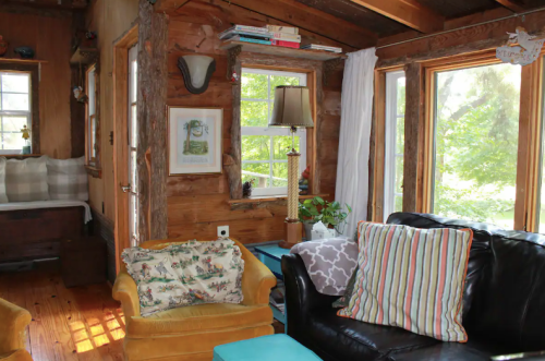 Cozy interior of a rustic cabin with wooden walls, a black couch, yellow armchair, and large windows letting in natural light.