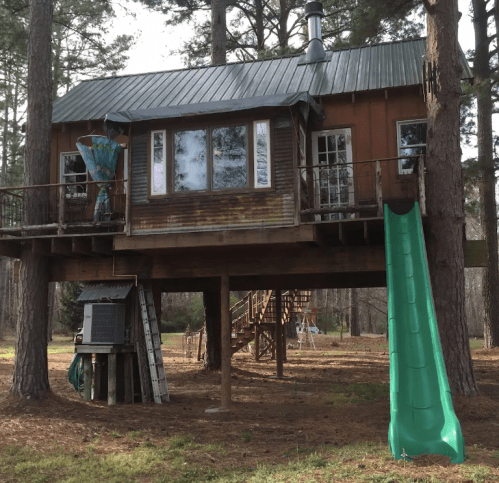 A wooden treehouse with a metal roof, featuring a slide and surrounded by tall trees in a grassy area.