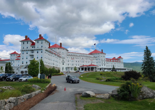 A large white hotel with red roofs, surrounded by greenery and mountains under a partly cloudy sky.