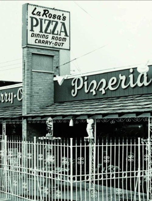 Black and white image of La Rosa's Pizzeria sign and exterior, featuring a fenced area and decorative statues.
