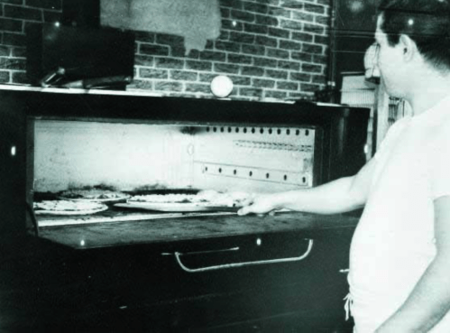A man using a pizza peel to remove pizzas from a brick oven in a vintage kitchen setting.