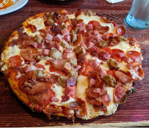 A large pizza topped with pepperoni, sausage, ham, and melted cheese on a wooden table.