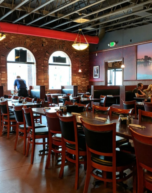 A spacious restaurant interior with wooden tables, chairs, and large windows, featuring a server and diners.