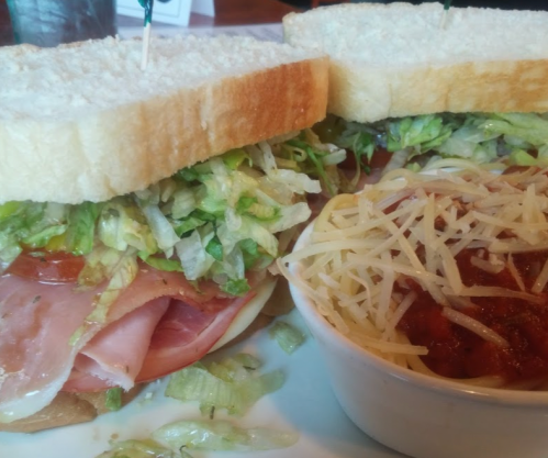 A close-up of a sandwich with ham, lettuce, and tomato, served with a side of marinara sauce and grated cheese.