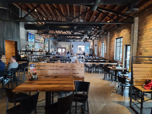 A spacious restaurant interior with wooden tables, black chairs, and a rustic design, featuring patrons dining and socializing.