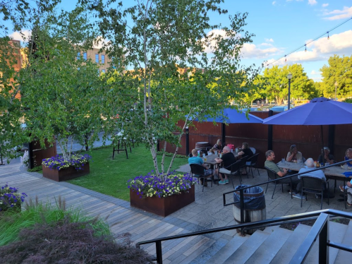 Outdoor dining area with tables under purple umbrellas, surrounded by greenery and flowers, on a sunny day.