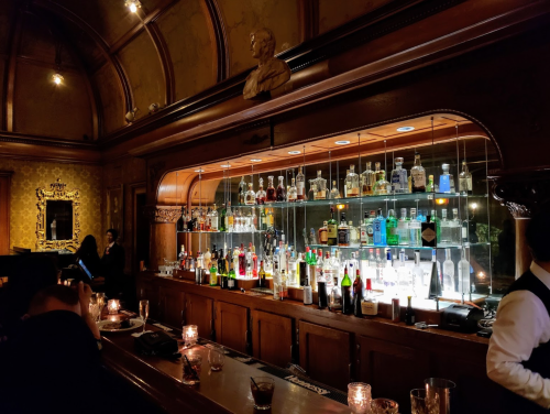 A dimly lit bar with a wooden counter, shelves lined with various bottles, and candles on the tables.