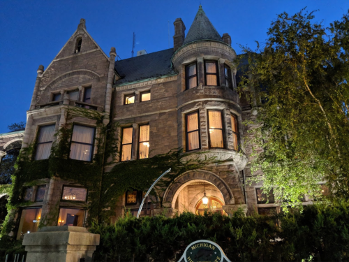A historic stone mansion with ivy, illuminated at dusk, surrounded by trees and a sign reading "Michigan."
